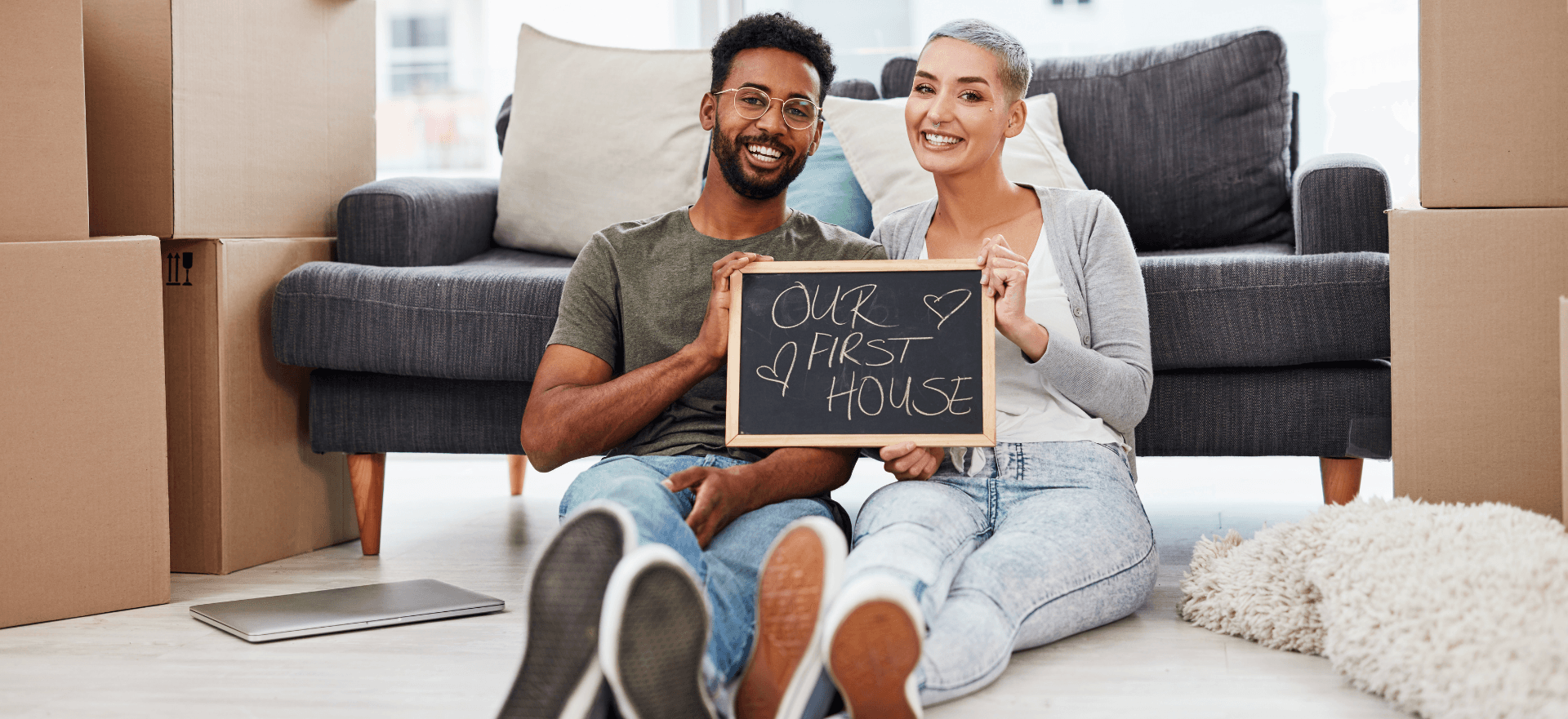 first-time-home-buyers-young-urban-couple-holding-a-sign-that-say-our-first-home-with-a-couch-and-moving-boxes-in-the-background
